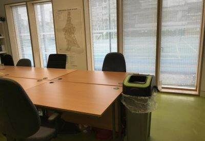 Office workspace with a cluster of wooden desks, black office chairs, and views through large windows.