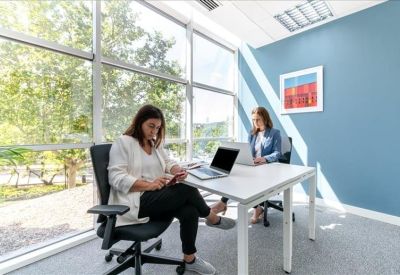 Bright corner office with two desks and large windows overlooking trees.