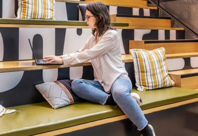 Worker using a laptop on tiered wooden seating with cushions and motivational wall text.