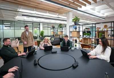 Team gathered around a large circular black desk in a modern coworking space with shelving units.