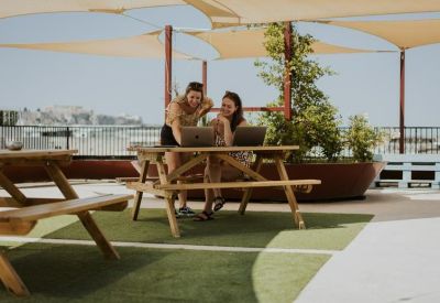 Rooftop terrace with people working at wooden picnic tables under sun sails.