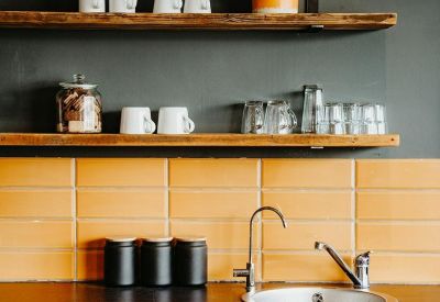 Communal kitchen area with orange tiled backsplash, wooden shelves, and light grey cabinets.
