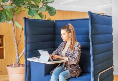 A woman working on a laptop in a private blue soundproof pod next to a large potted plant.