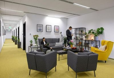 Bright communal lounge area with gray armchairs, a yellow wingback chair, and potted plants.