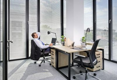 Bright corner office with two wooden desks, black ergonomic chairs, and floor-to-ceiling windows.