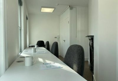 Long white desk in a bright office with several grey ergonomic chairs and a decorative fireplace.