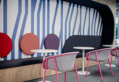 Communal breakout area featuring colorful wall cushions, small round tables, and pink-framed chairs.