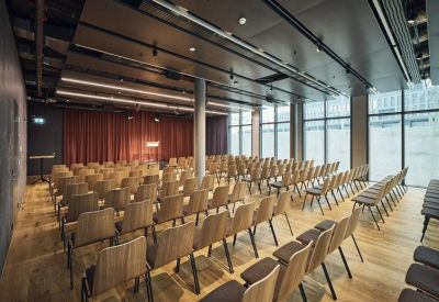 Large event hall arranged with rows of wooden chairs and a red stage curtain.