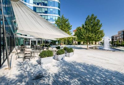 Sunlit outdoor patio with white shade sails, café seating, and a fountain.