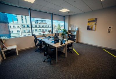 Open-plan office space with desks, ergonomic chairs, and large windows showing city views.