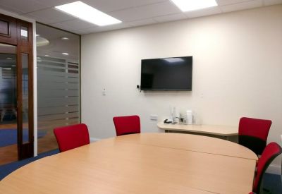 Modern meeting room with a round wooden table, red chairs, and a wall-mounted screen.