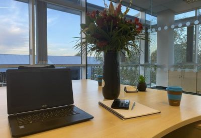 Close-up of a desk with a laptop, flowers in a vase, and a notepad by a window.