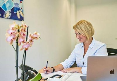 A woman working at a bright desk with a pink orchid and laptop.