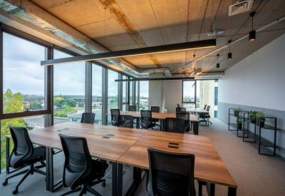 Open-plan office space featuring wooden desks, black chairs, and large windows with city views.