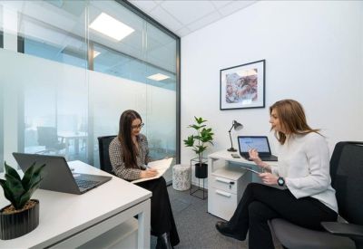 Two women discussing work in a modern private office with glass partitions.