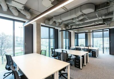 Open-plan office space with white desks and black ergonomic chairs under industrial ceilings.