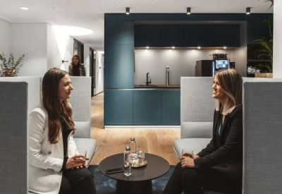 Two women talking in a communal lounge area with high-backed grey booths and a kitchen backdrop.