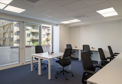 Spacious sunlit office with several white desks and ergonomic black chairs near large windows.