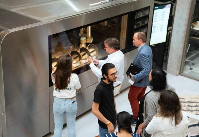 A group of people gathered around a large industrial lab machine in a modern atrium.