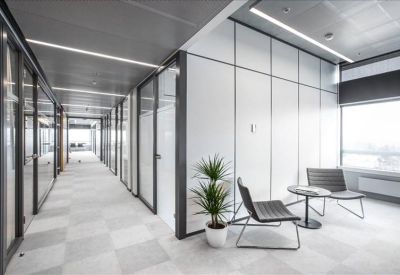 Bright modern office corridor with glass partitions and a small seating area with a plant.