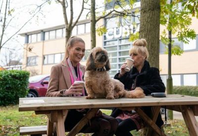 Two women with a dog sitting at an outdoor wooden picnic table with coffee.