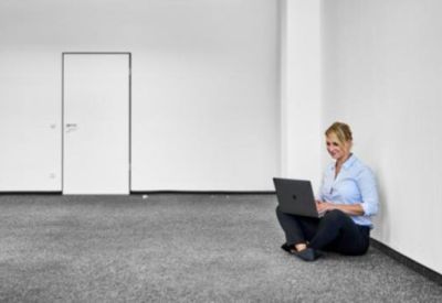 Minimalist, spacious white room with a professional woman sitting on the carpet floor using a laptop.