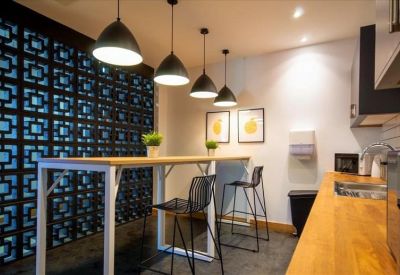 Modern communal kitchen area with high wooden table, stools, and pendant lighting.