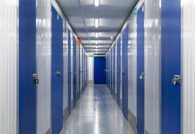 A long, brightly lit corridor of secure indoor self-storage units with blue doors.