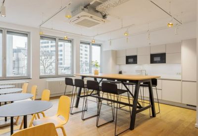 Bright office kitchen and dining area with a high wooden table and yellow chairs.