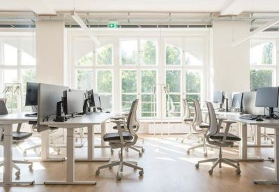 Bright open-plan office space featuring white sit-stand desks and ergonomic chairs near large windows.