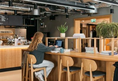 Modern communal cafe area with wooden counters, plants, and people working at long tables.