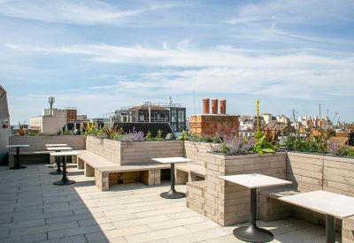 Sunny rooftop terrace with wooden planter benches, small white tables, and views over city rooftops.