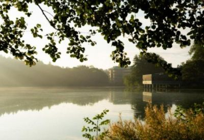 Exterior view of No. 1 Mereside, Alderley Park, overlooking a serene lake at sunrise with hanging tree branches.