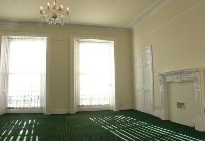 Bright room with green carpeting, two large sash windows, and a decorative white fireplace.