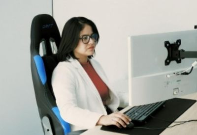 A woman working at a computer desk with a white blazer and black ergonomic chair.