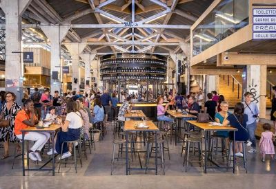 Lively communal dining hall with high ceilings, industrial trusses, and many people at wooden tables.