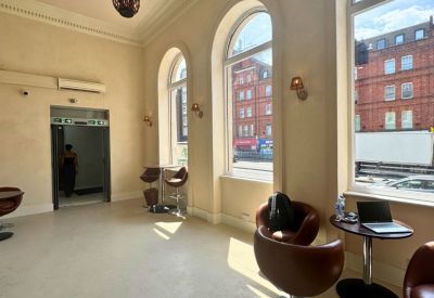 Sunlit communal lounge with high arched windows, brown leather chairs, and pendant lighting.