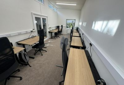 Bright open-plan office space featuring several long wooden desks and black ergonomic chairs.