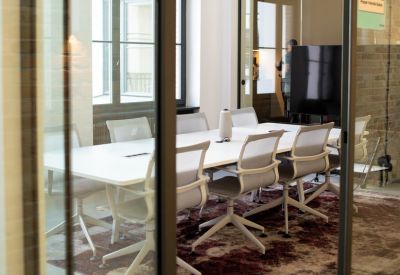 Meeting room with a long white table, grey chairs, and a decorative patterned rug behind glass walls.