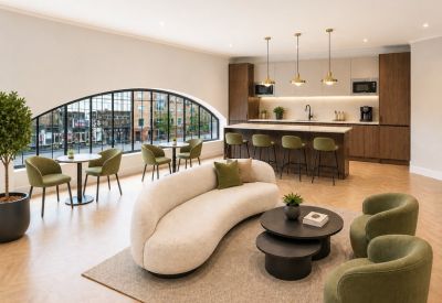 Bright communal lounge and kitchen area with a curved white sofa, green chairs, and arched window.