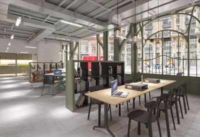 Bright coworking space with long wooden tables, books, and large industrial-style windows.