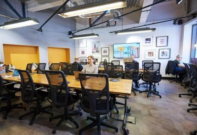Industrial-style open plan office with long wooden desks and black ergonomic chairs.