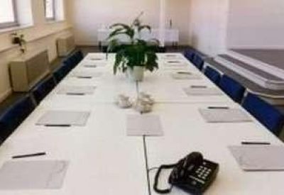 Spacious boardroom set up with a long white conference table, blue chairs, and a central plant.