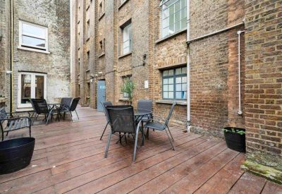Outdoor wooden deck terrace with black mesh chairs and tables between brick buildings.