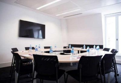 Bright conference room with a U-shaped table, black chairs, and a wall-mounted screen.