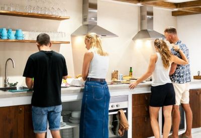 Modern shared kitchen with wooden cabinets, dual range hoods, and people preparing food.