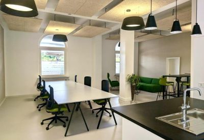 Spacious open-plan office with long white desks, pendant lighting, and a kitchen area.
