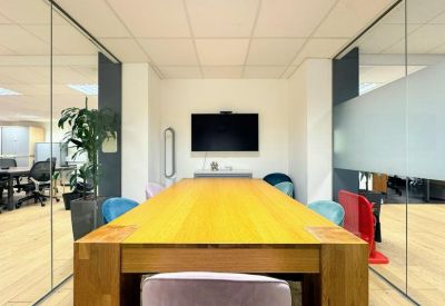 Sunlit meeting room with a large wooden table, colourful seating, and glass partitions.