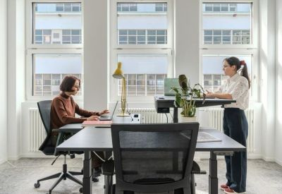 Two people working at a shared black height-adjustable desk in a bright office with large windows.