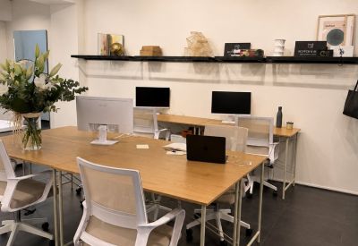 Bright office workspace featuring light wood desks, white ergonomic chairs, and wall-mounted shelving.